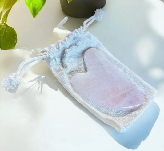 Rose quartz gua sha glow stone in a white drawstring bag on a light surface with a plant in the background.